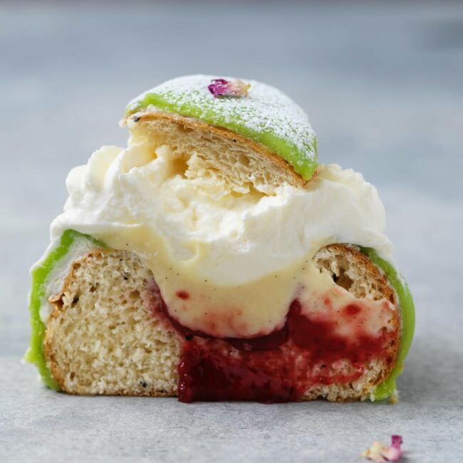 Närbild av en prinsess-semla med grön marsipan, fluffig grädde, len vaniljkräm och hallonsylt.